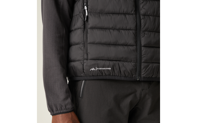 Nahaufnahme des Oberkörpers mit schwarzer gesteppter Weste mit Reißverschluss, seitlicher Reißverschlusstasche und weißem "FEATHER FREE"-Logo am Saum; dunkles Langarmshirt, dunkle Hose und sichtbare linke Hand