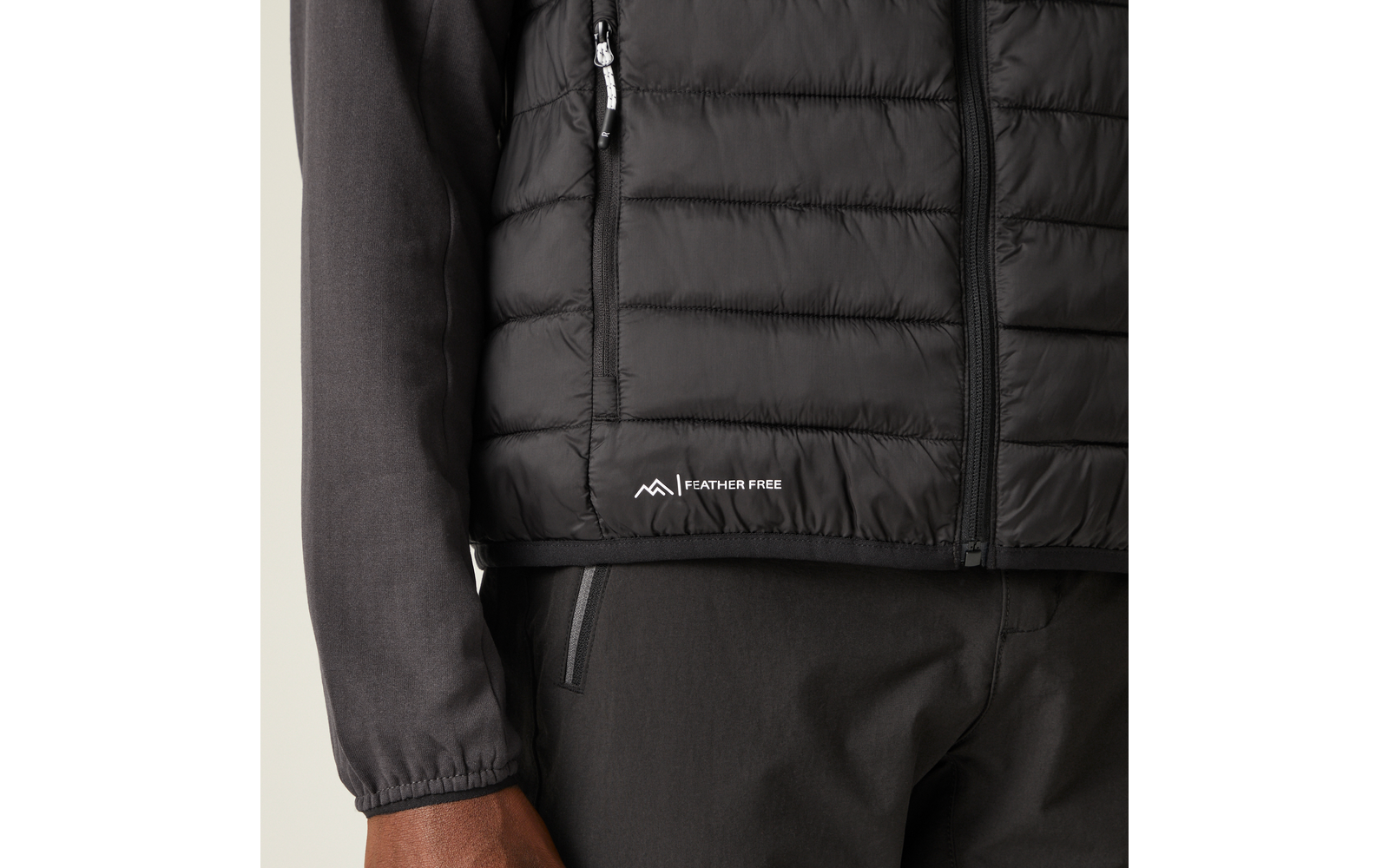 Nahaufnahme des Oberkörpers mit schwarzer gesteppter Weste mit Reißverschluss, seitlicher Reißverschlusstasche und weißem "FEATHER FREE"-Logo am Saum; dunkles Langarmshirt, dunkle Hose und sichtbare linke Hand