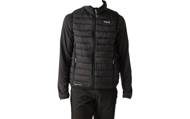 Persona che indossa un gilet imbottito nero sopra una maglia nera a maniche lunghe e pantaloni neri, in piedi davanti a uno sfondo chiaro uniforme, guarda verso sinistra, mani lungo i fianchi