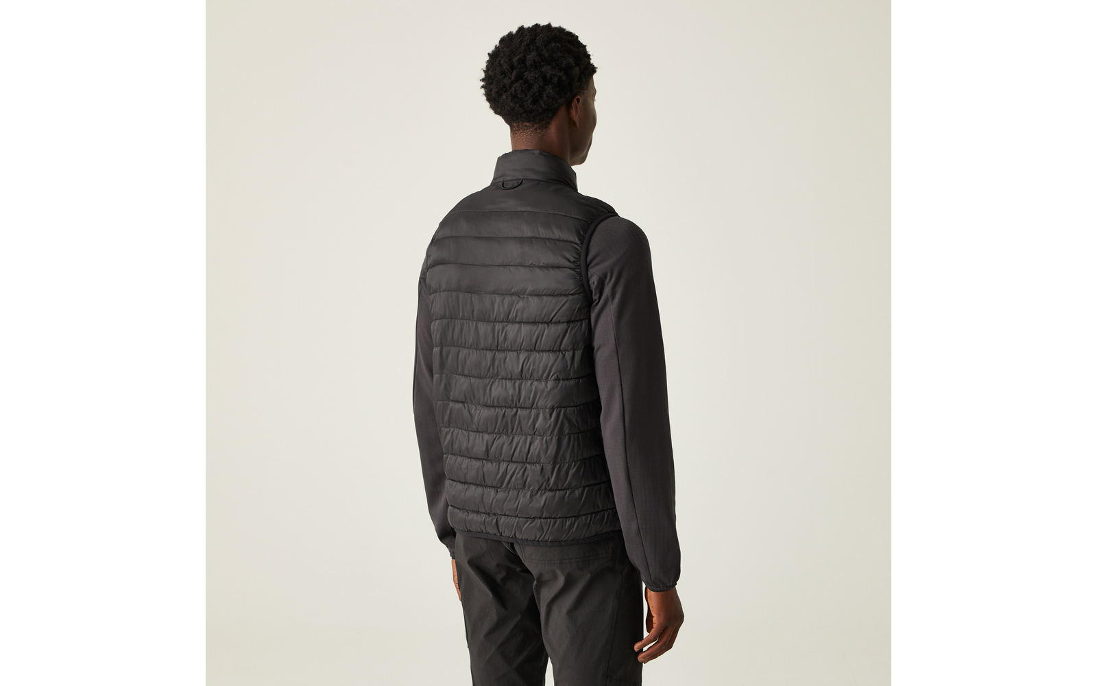 Person von hinten, schwarze gesteppte Weste über langärmligem schwarzen Oberteil und dunkle Hose, heller Hintergrund