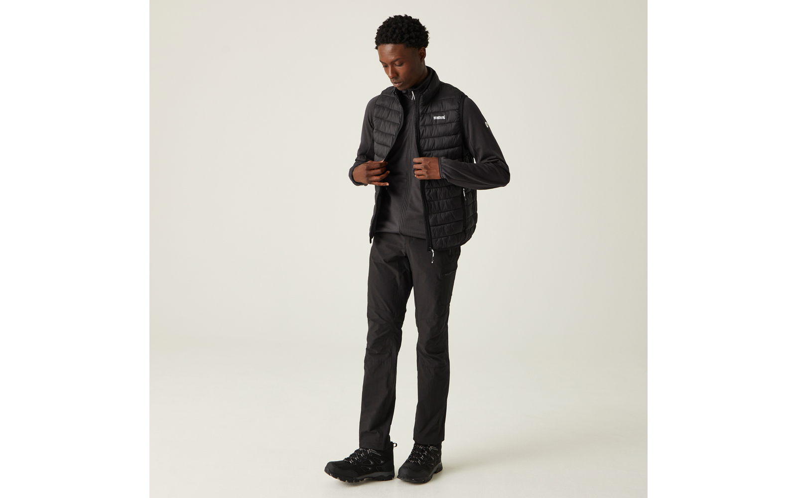 Stehende Person vor hellem Hintergrund, trägt schwarze gesteppte Weste über schwarzem Langarmshirt, schwarze Hose und schwarze Wanderschuhe, Blick nach unten, rechte Hand am Reißverschluss der Weste