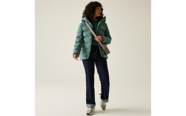 Frau mit lockigem Haar trägt eine grüne Steppjacke, dunkle Hose und weiße Schuhe, sie hält eine braune Tasche auf der Schulter