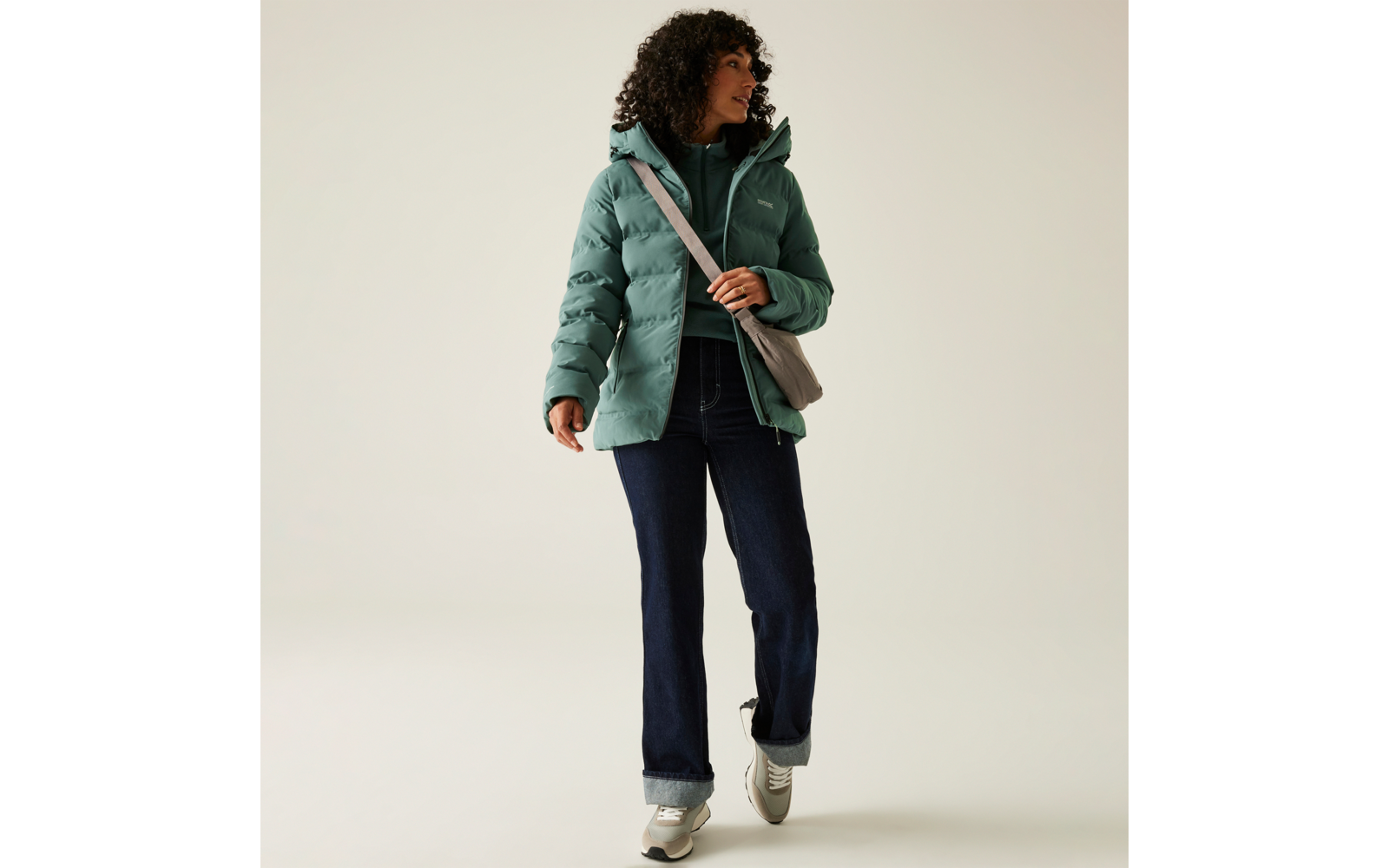 Frau mit lockigem Haar trägt eine grüne Steppjacke, dunkle Hose und weiße Schuhe, sie hält eine braune Tasche auf der Schulter