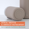 Zwei beige zylindrische Becher auf weißer Fläche, einer liegt auf der Seite und zeigt den runden Boden mit geprägtem Logo; orangefarbener Banner mit weißer und grüner Schrift unten links