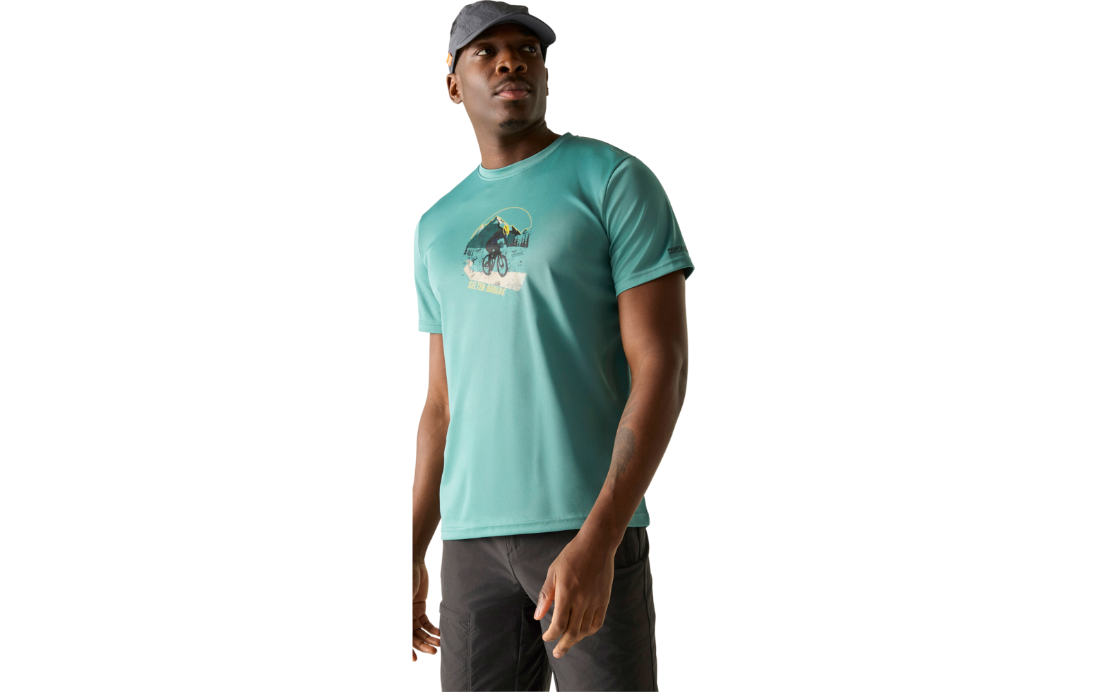 Mann steht leicht zur Seite, trägt türkisfarbenes T‑Shirt mit Grafik (Fahrrad und Berge), dunkle Kappe und Shorts, heller Hintergrund