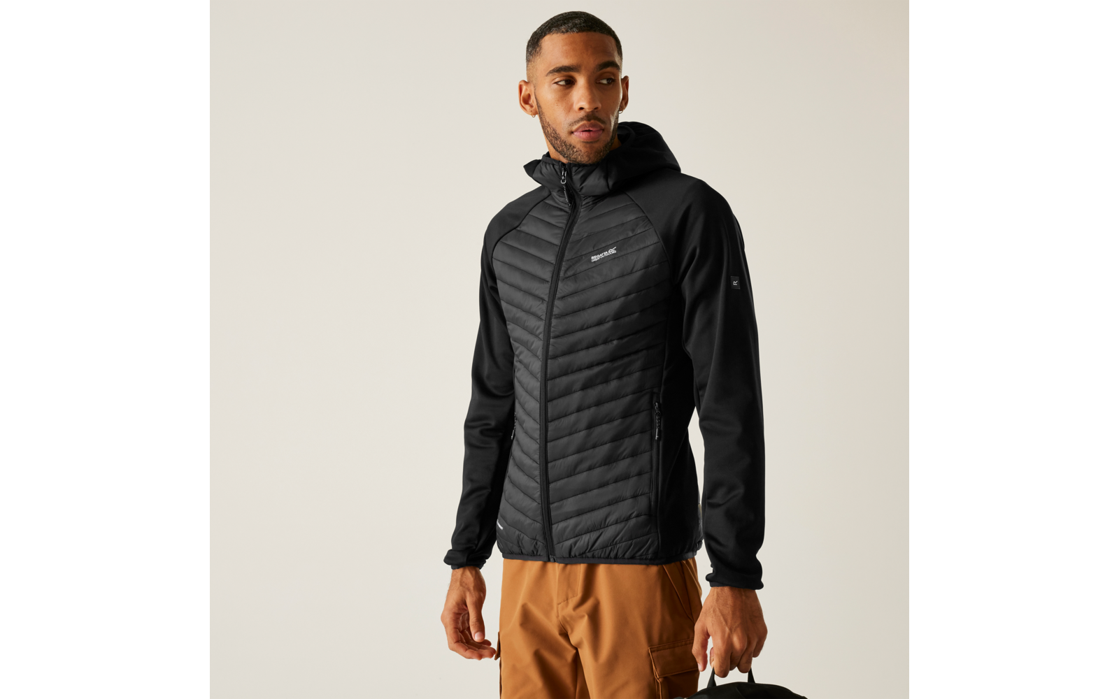 Person trägt schwarze gesteppte Kapuzenjacke mit Reißverschluss, schwarze Ärmel, kleines Logo auf der Brust, braune Hose, hält eine schwarze Tasche in der rechten Hand, heller Hintergrund