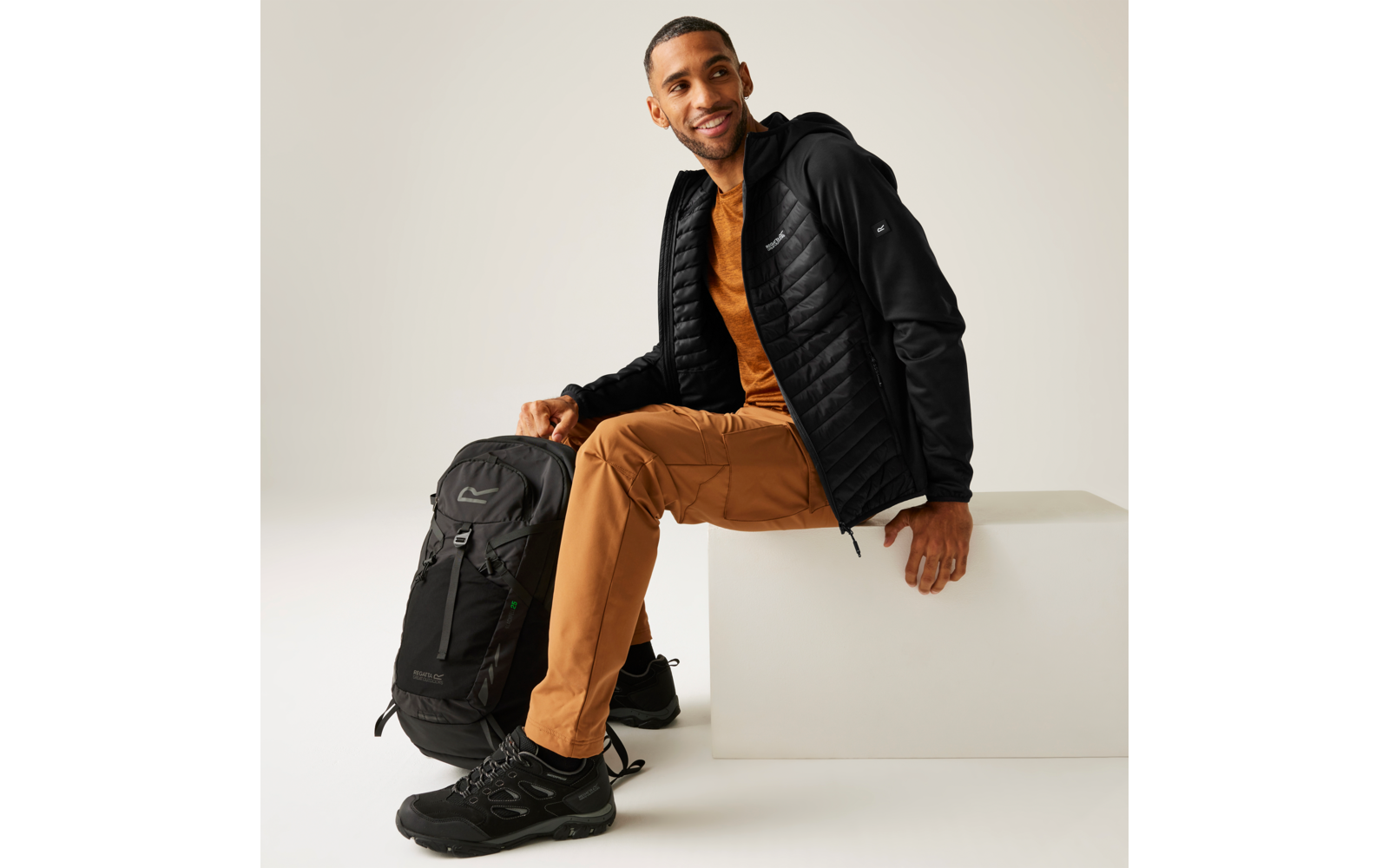 Person sitzt auf weißem Block, trägt schwarze gesteppte Jacke mit Kapuze, braune Hose und schwarze Schuhe; schwarzer Rucksack steht neben der Person auf dem Boden