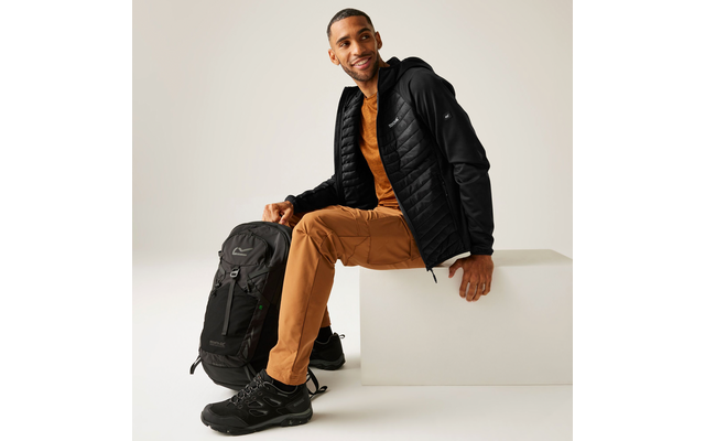 Mann sitzt auf einer weißen Fläche, trägt eine schwarze Jacke, braune Hose und schwarze Schuhe, neben ihm steht ein schwarzer Rucksack