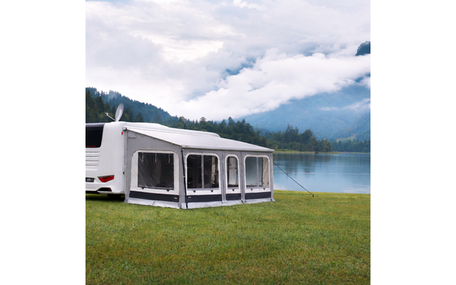 Weißes Wohnmobil mit grauem Vorzelt auf Gras am Ufer eines ruhigen Sees; bewaldete Berge und niedrige Wolken im Hintergrund