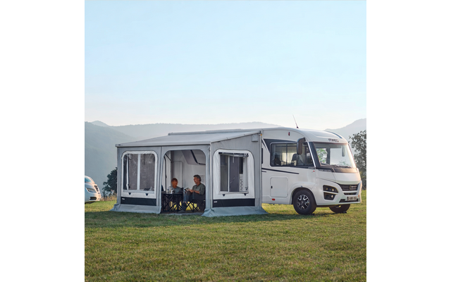 Wohnmobil mit angebautem Vorzelt, zwei Personen sitzen am Tisch, Wiese und Hügel im Hintergrund