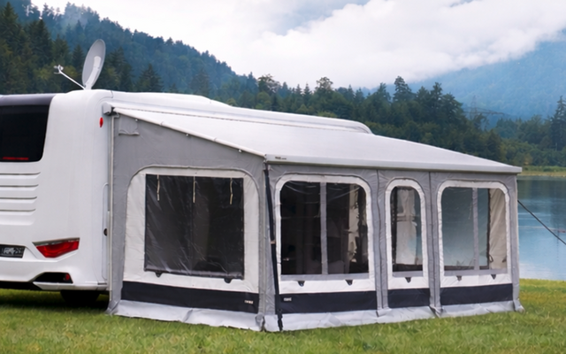 Weißes Wohnmobil mit grauem Vorzelt auf Gras am Ufer eines ruhigen Sees; bewaldete Berge und niedrige Wolken im Hintergrund