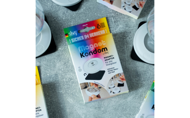 Verpackung eines Magnet-Kondoms auf grauem Untergrund, mit weißen Tassen und Tellern im Hintergrund