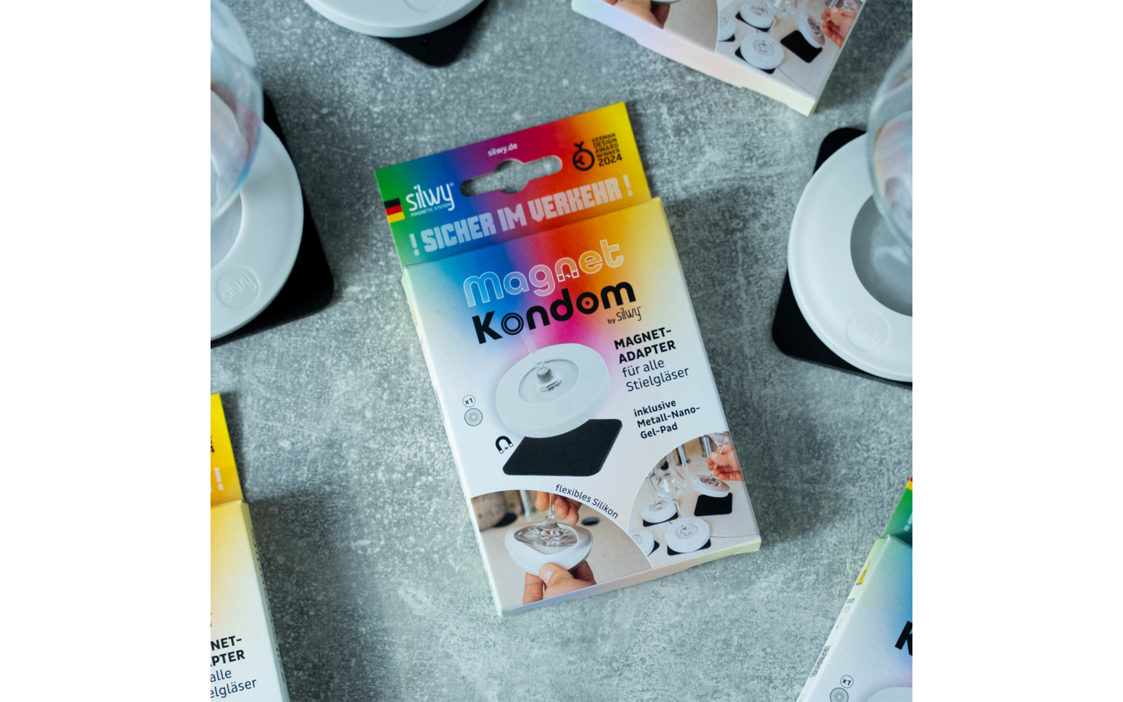 Verpackung eines Magnet-Kondoms auf grauem Untergrund, mit weißen Tassen und Tellern im Hintergrund