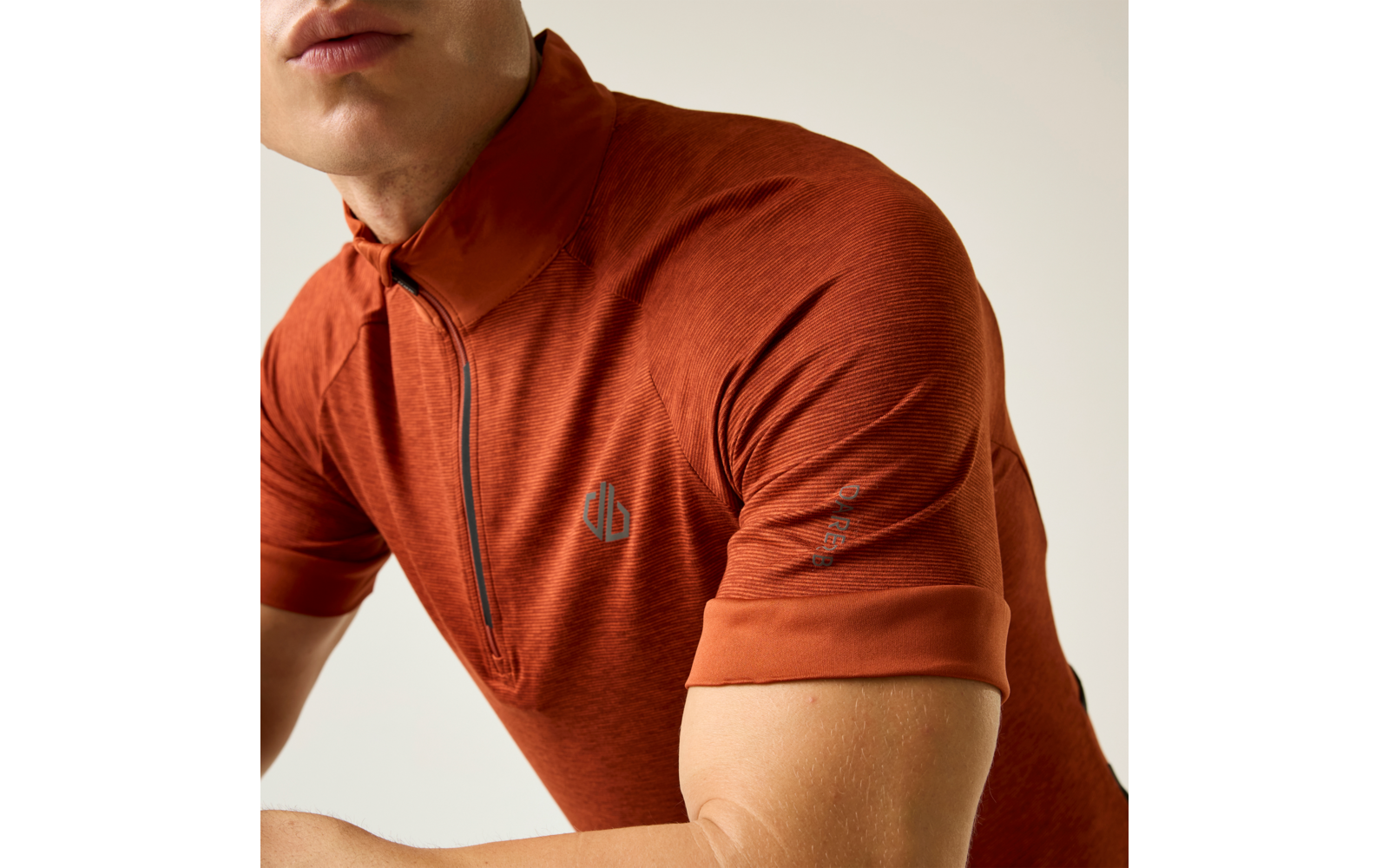 Oberkörper einer Person in orangefarbenem, kurzärmeligem Polo mit kurzem Reißverschluss; Unterkiefer sichtbar, verschränkte Arme, heller Hintergrund