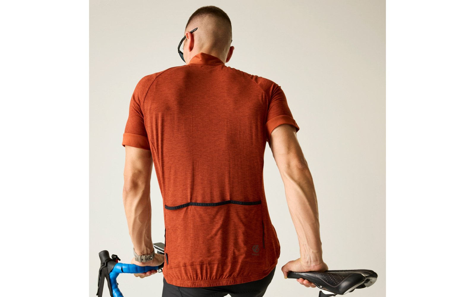 Person von hinten, trägt orangefarbenes kurzärmeliges Radtrikot, hält mit linker Hand einen blauen Lenker und mit rechter Hand einen schwarzen Fahrradsattel, hell einfarbiger Hintergrund