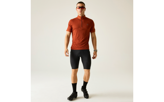 Stehende Person in Ganzkörperaufnahme vor hellem einfarbigem Hintergrund, trägt rotes kurzärmeliges Shirt mit kurzem Reißverschluss, enge schwarze Shorts, schwarze Socken, schwarze Schuhe, Sonnenbrille und Armbanduhr