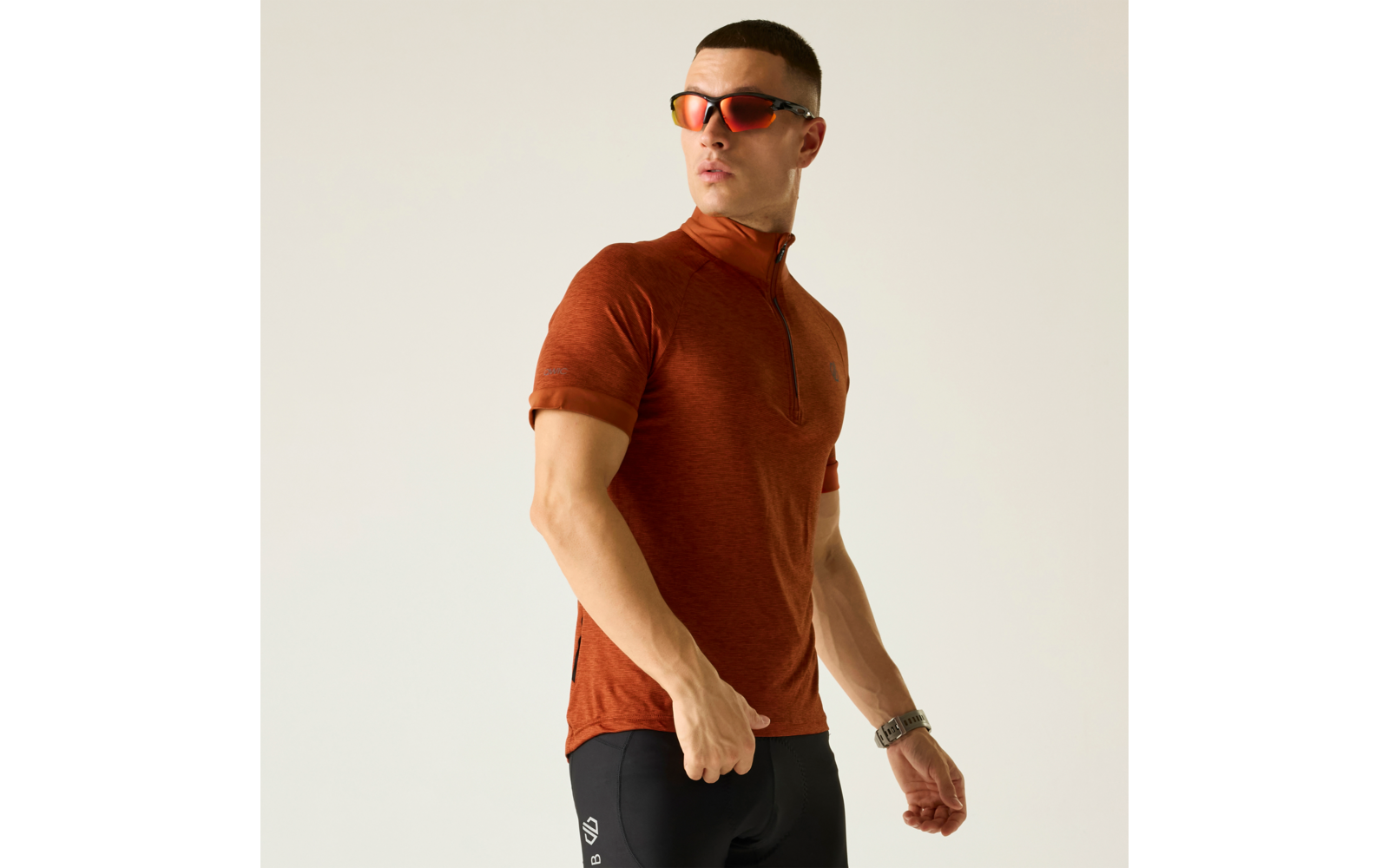 Person in orangefarbenem kurzärmligem Zip-Top, schwarzen engen Shorts mit weißem Logo am Oberschenkel, roten verspiegelten Sonnenbrillen und Armbanduhr, steht vor hellem Hintergrund und schaut zur Seite