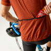 Person von hinten in rostfarbenem Trikot steckt ein Energiegel in die Rückentasche und hält einen Fahrradhelm