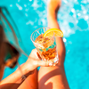 Beine und eine Hand, die ein facettiertes Glas mit eisigem, bernsteinfarbenem Getränk und einer Zitronenscheibe am Rand hält; türkisfarbenes Poolwasser im Hintergrund