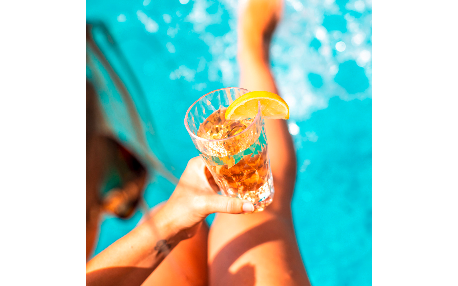 Beine und eine Hand, die ein facettiertes Glas mit eisigem, bernsteinfarbenem Getränk und einer Zitronenscheibe am Rand hält; türkisfarbenes Poolwasser im Hintergrund
