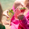 Zwei Hände halten Gläser: links klares Glas mit Eis, Limette und Minze; rechts Glas mit rosa Getränk, Strohhalm und Wassermelonenscheibe am Rand; sichtbare Schultern mit rosa Kleidung; unscharfer Wasserhintergrund