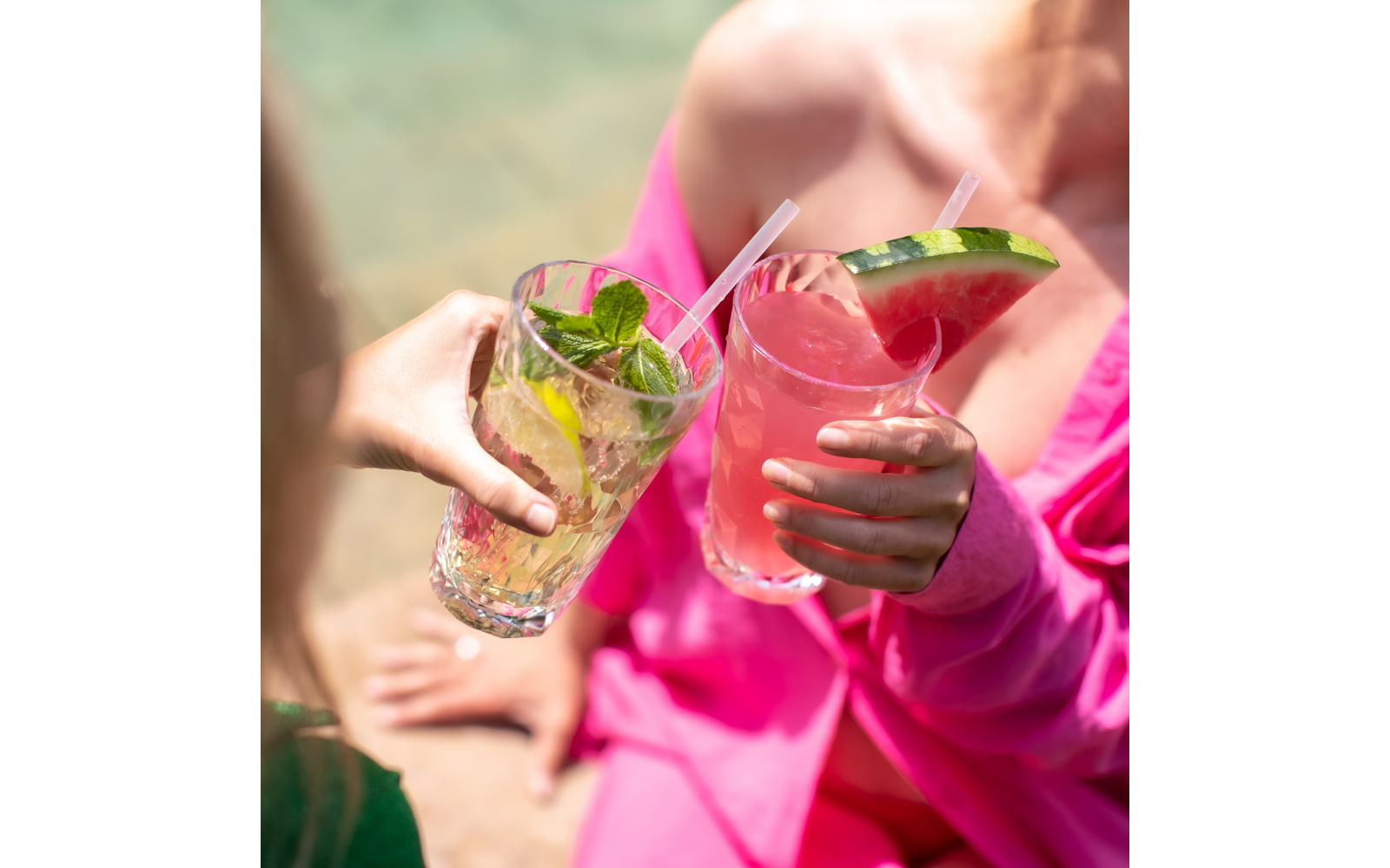 Zwei Hände halten Gläser: links klares Glas mit Eis, Limette und Minze; rechts Glas mit rosa Getränk, Strohhalm und Wassermelonenscheibe am Rand; sichtbare Schultern mit rosa Kleidung; unscharfer Wasserhintergrund