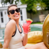 Lächelnde Frau mit Sonnenbrille und weißem Badeanzug sitzt auf goldener Luftmatratze am Pool und hält ein rosa Getränk mit einer Scheibe Wassermelone