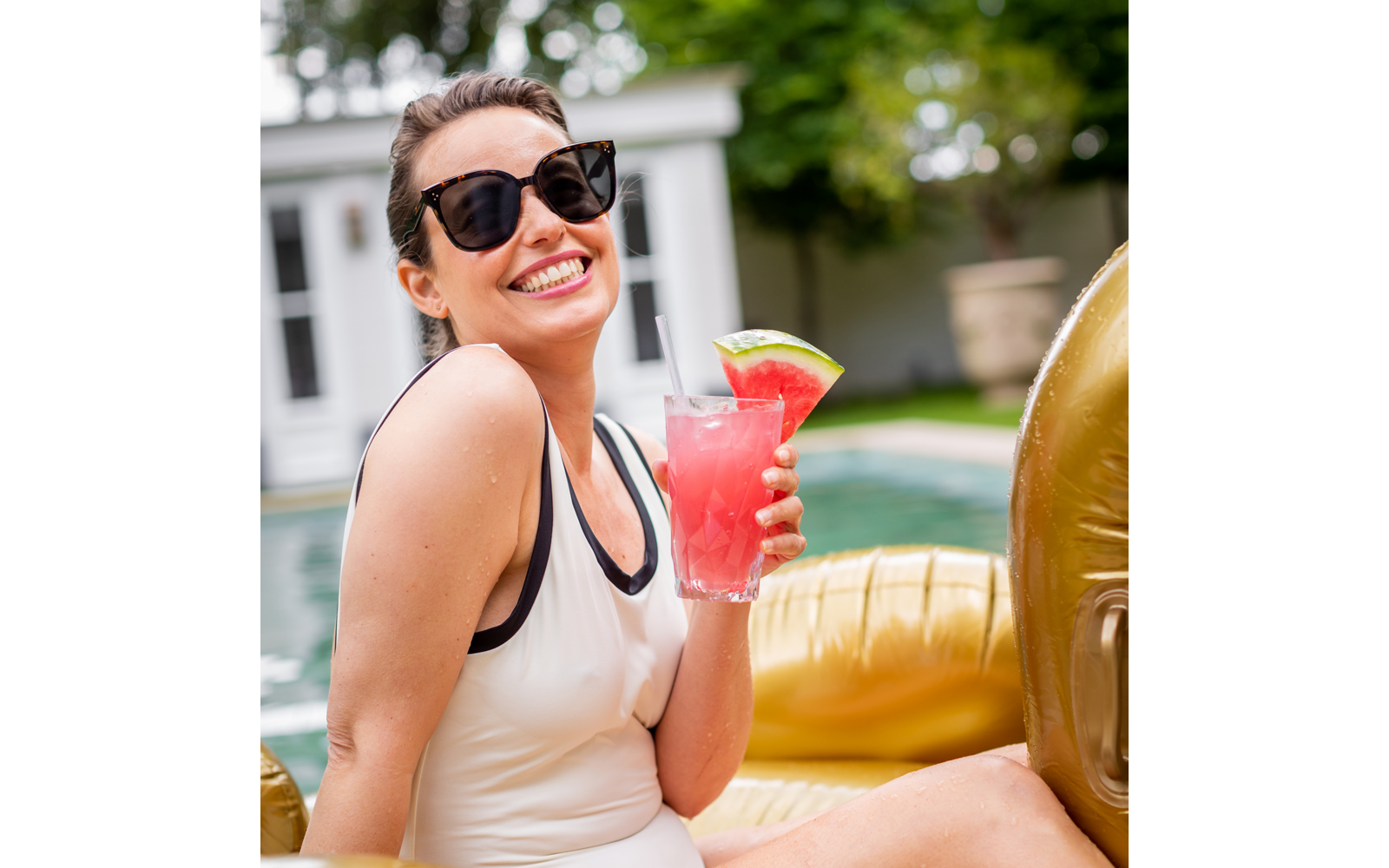 Lächelnde Frau mit Sonnenbrille und weißem Badeanzug sitzt auf goldener Luftmatratze am Pool und hält ein rosa Getränk mit einer Scheibe Wassermelone