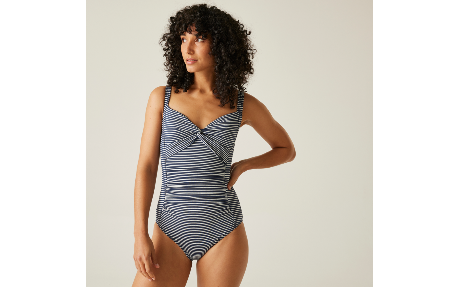 Femme aux cheveux bouclés foncés portant un maillot de bain une-pièce rayé bleu et blanc, une main sur la hanche, regard vers la gauche, fond clair uni