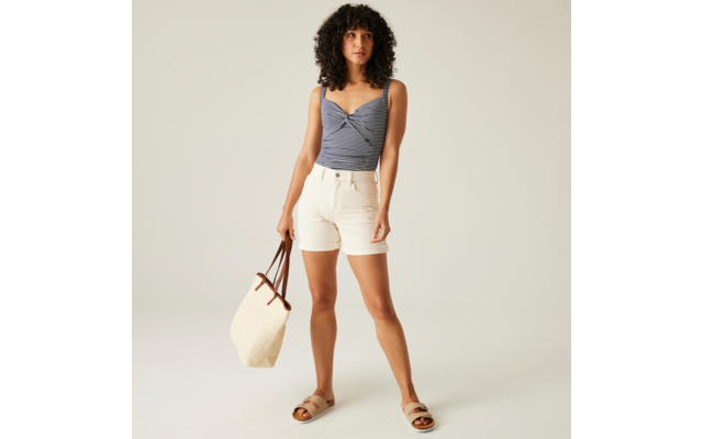 Personne aux cheveux bouclés mi-longs debout, portant un haut bleu avec un détail torsadé à l'avant, un short blanc, des sandales et tenant un sac en tissu clair sur fond clair