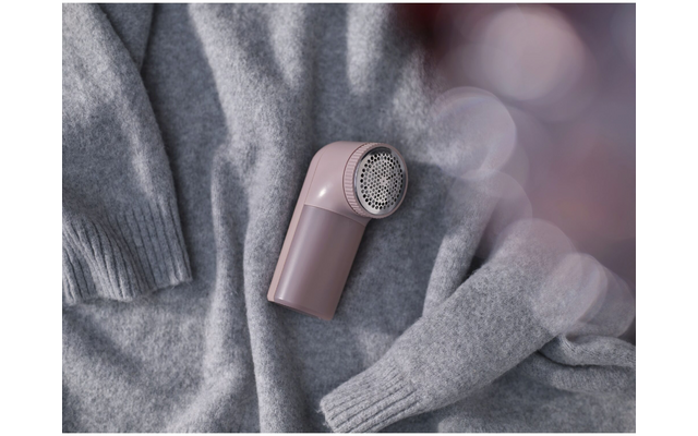 Kleines rosafarbenes Handgerät mit rundem Metallgitter liegt auf einem grauen Strickpullover, Ärmel sichtbar