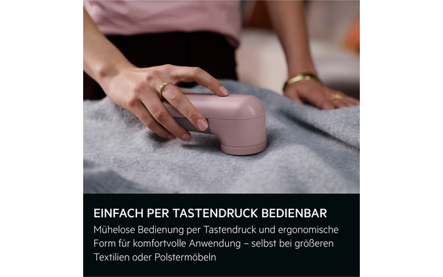 Hand mit goldenen Ringen hält ein rosafarbenes, rundes Handgerät auf grauem Stoff; rosa Oberteil im Hintergrund