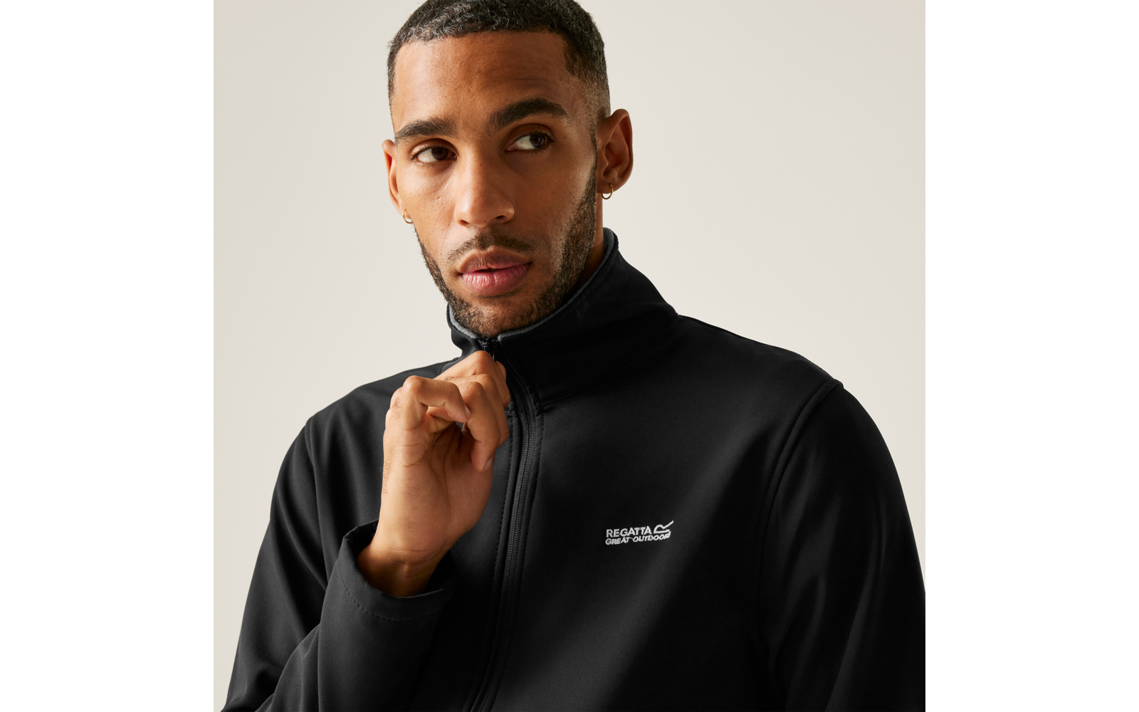 Close-up van een persoon die aan de ritssluiting van een zwarte jas met wit borstlogo trekt; kort haar, oorbel, baard, kijkt naar rechts, lichte achtergrond