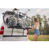 Femme en haut sans manches et short en jean fixe un vélo sur le porte-vélos arrière d'un camping-car blanc sur une prairie avec des arbres en arrière-plan