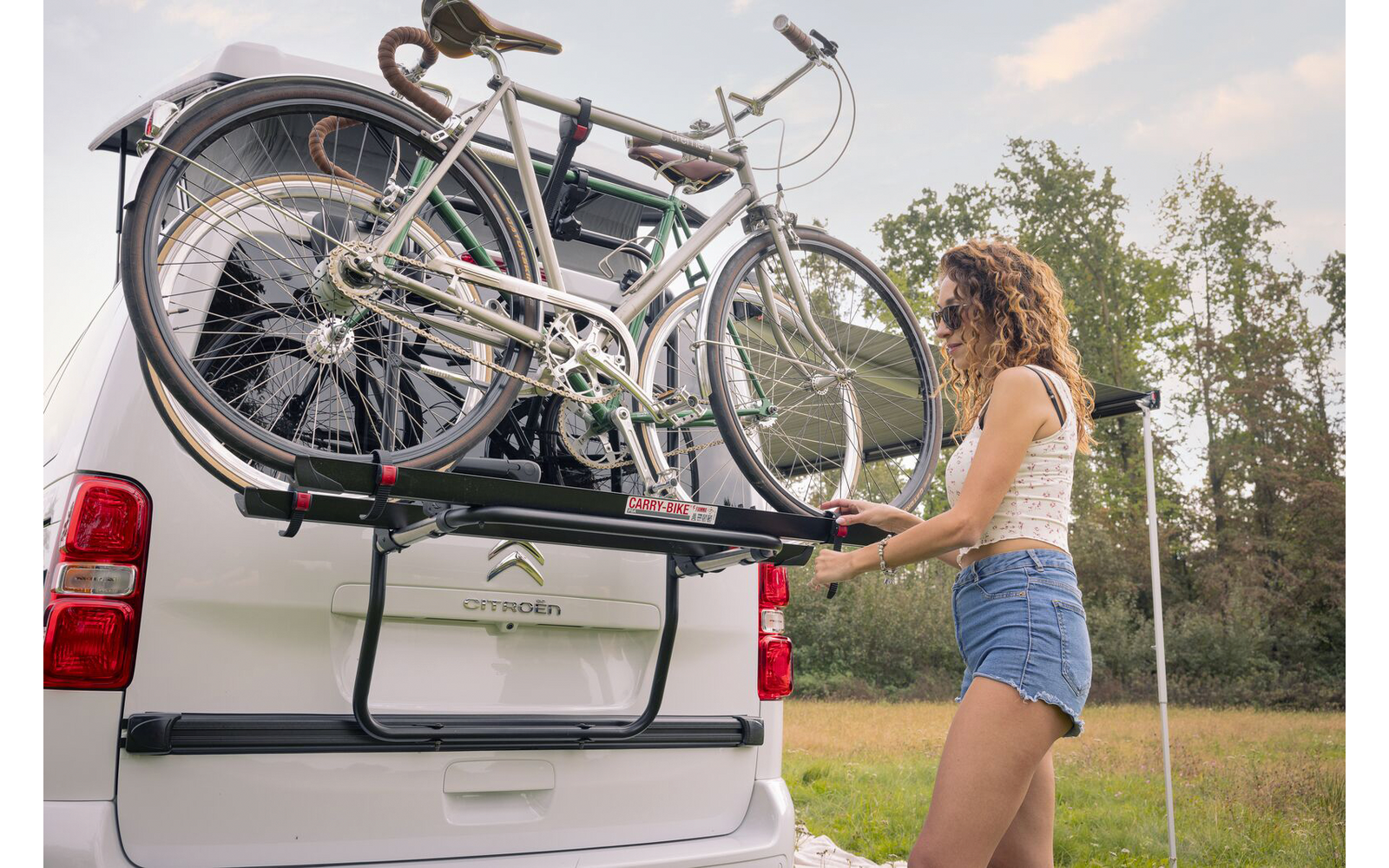 Femme en haut sans manches et short en jean fixe un vélo sur le porte-vélos arrière d'un camping-car blanc sur une prairie avec des arbres en arrière-plan