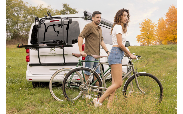 Un homme et une femme sont debout avec deux vélos à côté d'une camionnette blanche sur une prairie, arbres d'automne en arrière-plan