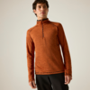 Un homme porte un pull orange à manches longues avec une fermeture éclair jusqu'à la poitrine et un pantalon noir devant un fond uni et clair