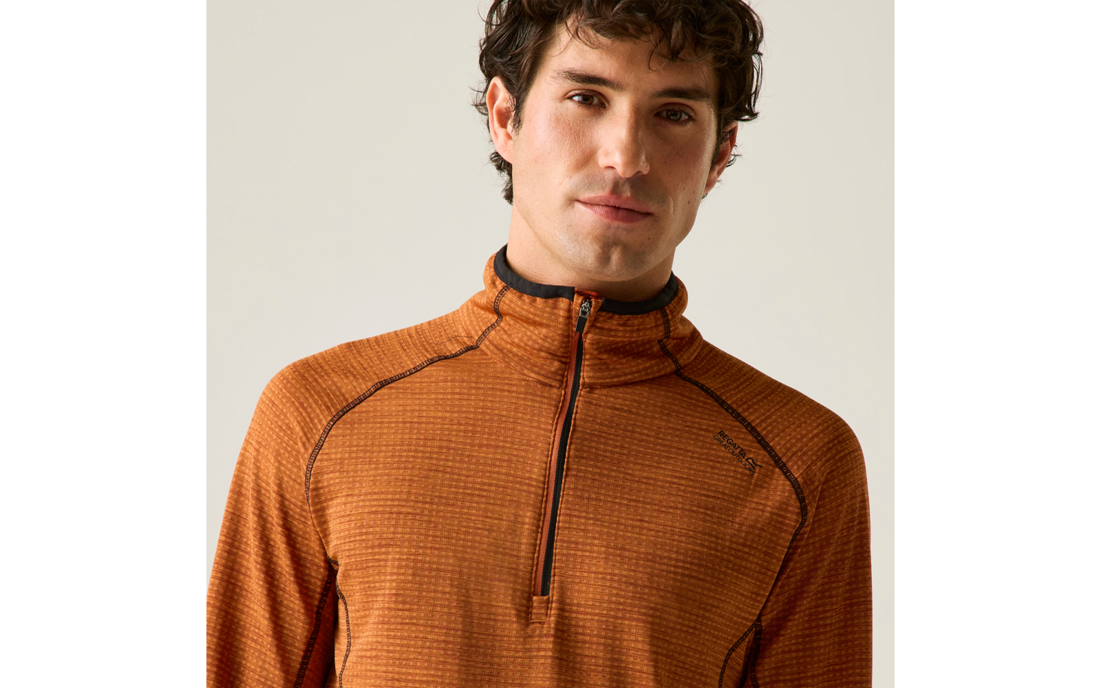 Personne aux cheveux bruns et bouclés, portant un haut de sport orange texturé avec une courte fermeture éclair, fond uni lumineux