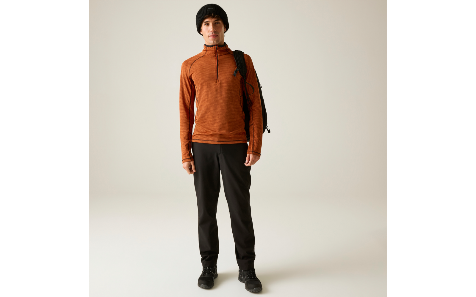 Personne portant un haut à manches longues orange, un bonnet noir, un pantalon noir et un sac à dos, debout devant un fond clair