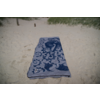 Serviette de plage rectangulaire à motif ornemental bleu et blanc posée à plat sur du sable clair, herbe de dunes et deux pieux en bois en arrière-plan
