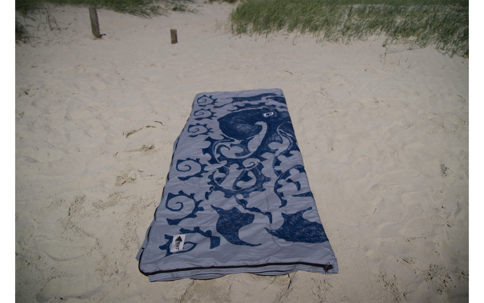 Serviette de plage rectangulaire à motif ornemental bleu et blanc posée à plat sur du sable clair, herbe de dunes et deux pieux en bois en arrière-plan
