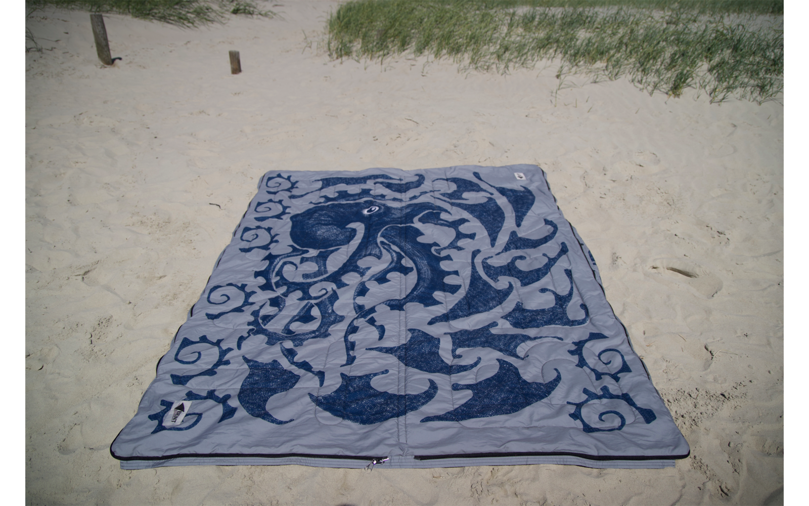 Serviette de plage rectangulaire bleue à motif, posée à plat sur du sable clair; herbe des dunes en haut