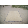 Serviette de plage rectangulaire beige posée à plat sur du sable clair; herbe de dune et deux petits poteaux en bois en arrière-plan
