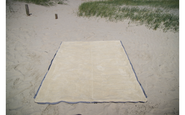 Serviette de plage rectangulaire beige posée à plat sur du sable clair; herbe de dune et deux petits poteaux en bois en arrière-plan