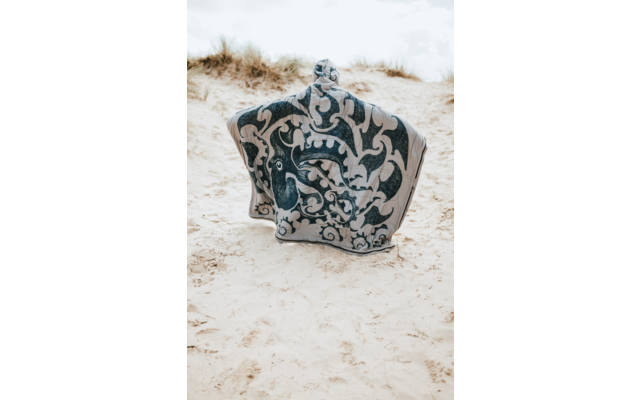 Objet à motif bleu et blanc à sommet arrondi posé sur du sable clair, dunes avec herbe sèche et ciel nuageux en arrière-plan