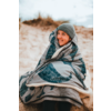 Personne assise enveloppée dans une grosse couverture sur une dune de sable, portant un bonnet gris en tricot et souriant