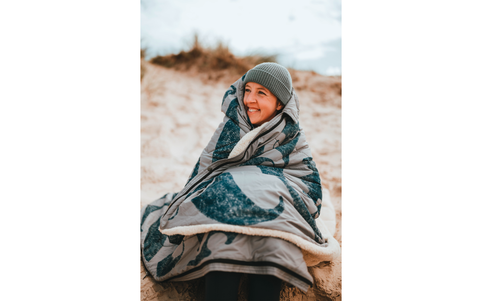 Personne assise enveloppée dans une grosse couverture sur une dune de sable, portant un bonnet gris en tricot et souriant