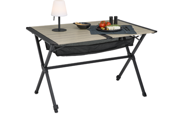 Tisch mit heller Holzplatte und schwarzem Metallgestell, schwarzes Netzfach darunter. Auf der Platte eine Tischlampe, eine Schale mit Holzlöffel, ein dunkles Tuch und ein Teller mit roten Tomaten und weißen Scheiben