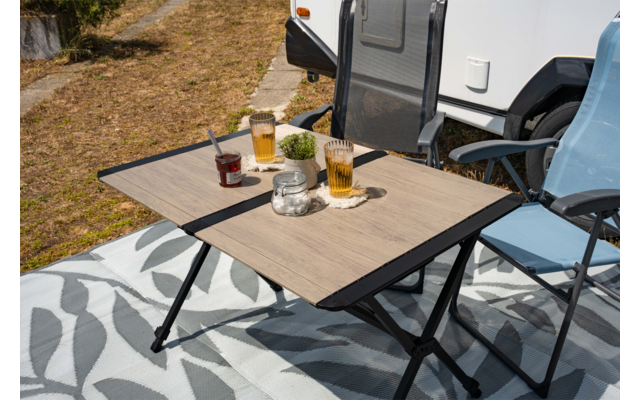 Klapptisch mit zwei hohen Gläsern mit bernsteinfarbener Flüssigkeit und Schaum, Marmeladenglas mit Löffel, kleine Dose, Topf mit Pflanze, zwei Campingklappstühle neben einem Wohnmobil auf einer gemusterten Decke