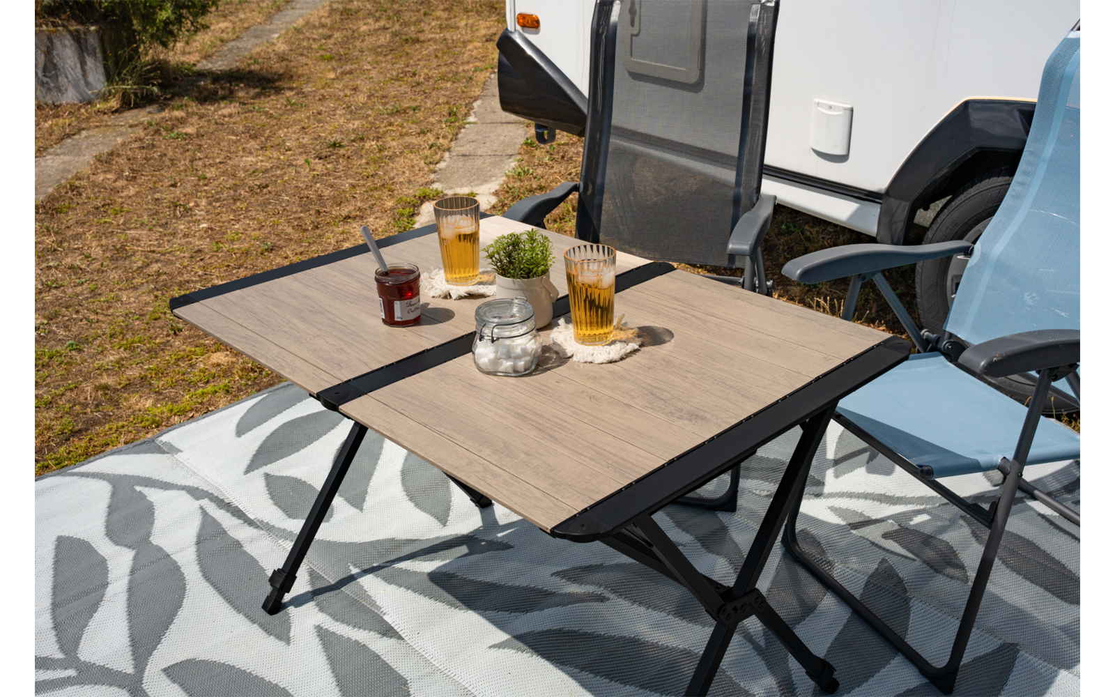 Klapptisch mit zwei hohen Gläsern mit bernsteinfarbener Flüssigkeit und Schaum, Marmeladenglas mit Löffel, kleine Dose, Topf mit Pflanze, zwei Campingklappstühle neben einem Wohnmobil auf einer gemusterten Decke